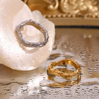 The gold and silver rings on a wet, reflective surface next to a seashell, showing their waterproof quality.