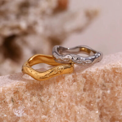 The silver wavy ring is stacked on top of the gold wavy ring while resting on a natural stone surface.