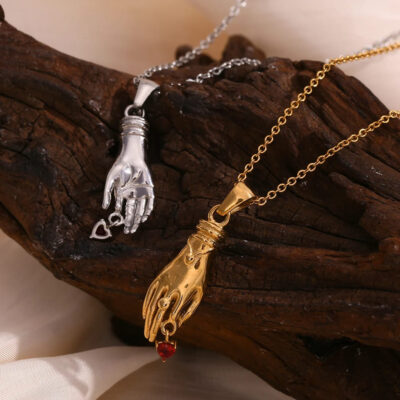 Close up of the Victorian-style hand pendant necklaces in both 18K gold and polished silver, resting on dark wood. Each features a dangling red heart charm.